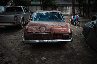 An old, rusted car parked in an outdoor lot shows significant deterioration, with paint peeling off the rear. A pickup truck is parked to the left, and a colorful scooter is visible in the background, parked near a concrete wall.