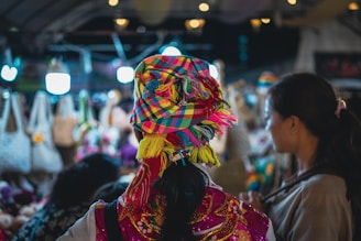 A bustling market scene with colorful textiles and clothing. The central figure is wearing a vibrant, multi-colored headscarf and traditional dress. Surrounding them are various handcrafted items, including bags, displayed under warm, illuminated lights.