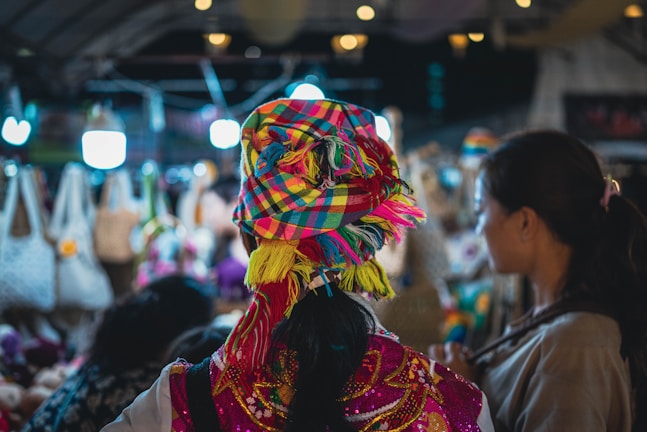 A bustling market scene with colorful textiles and clothing. The central figure is wearing a vibrant, multi-colored headscarf and traditional dress. Surrounding them are various handcrafted items, including bags, displayed under warm, illuminated lights.