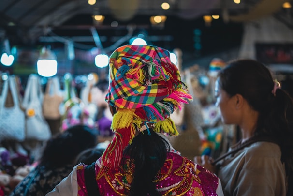 A bustling market scene with colorful textiles and clothing. The central figure is wearing a vibrant, multi-colored headscarf and traditional dress. Surrounding them are various handcrafted items, including bags, displayed under warm, illuminated lights.