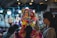 A bustling market scene with colorful textiles and clothing. The central figure is wearing a vibrant, multi-colored headscarf and traditional dress. Surrounding them are various handcrafted items, including bags, displayed under warm, illuminated lights.