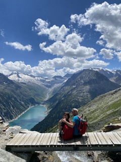 A couple with backpacks sits on a wooden bridge overlooking a breathtaking mountain landscape. Below them, a river flows into a turquoise lake surrounded by lush green hills and towering peaks. Puffy white clouds dot the bright blue sky, creating a serene and picturesque scene.
