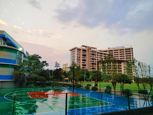 A modern university campus features a large multi-story building labeled 'Daffodil International University,' surrounded by greenery and a well-maintained basketball court. The court is wet, indicating recent rain, and there are trees and landscaped bushes around the area. The sky is overcast with hints of sunlight breaking through the clouds.