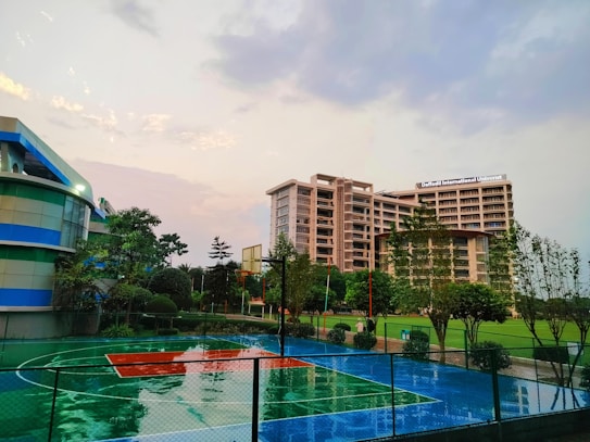 A modern university campus features a large multi-story building labeled 'Daffodil International University,' surrounded by greenery and a well-maintained basketball court. The court is wet, indicating recent rain, and there are trees and landscaped bushes around the area. The sky is overcast with hints of sunlight breaking through the clouds.