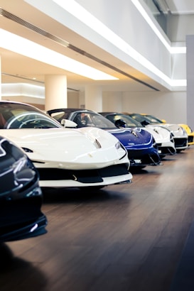 Displayed in a showroom, several luxury sports cars are lined up in a row on a polished wooden floor. The cars are primarily in colors like white, blue, black, and yellow, and are parked under modern, bright lighting, reflecting a clean and pristine environment.