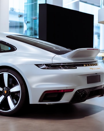 The image depicts the rear view of a sleek, white sports car displayed indoors. The car features a prominent spoiler and stylish rear lights. The Porsche logo is visible, along with dual exhausts at the lower bumper. The backdrop shows a modern, clean setting with glass elements and ambient lighting.