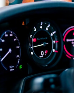 A close-up view of a car dashboard featuring several analog gauge displays, including a speedometer, tachometer, and fuel gauge. The displays are illuminated with different colored lights, including white, red, and green indicators. There are visible numbers and needle pointers, along with warning symbols and text.