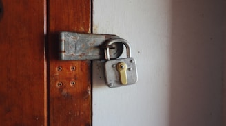 A robust silver padlock is securely fastened to a heavily weathered metal latch on a wooden door. The wooden surface shows signs of age with its faded brown color and visible screw holes.