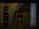 A dimly lit room with visible bunk beds, shelving units, and a washbasin. The interior appears sparse and functional, with a simple setup for personal items. The presence of folded towels and toiletries indicates a living space.