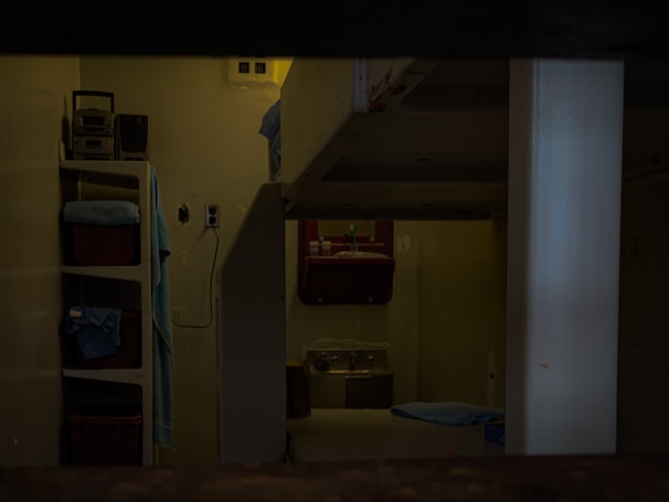 A dimly lit room with visible bunk beds, shelving units, and a washbasin. The interior appears sparse and functional, with a simple setup for personal items. The presence of folded towels and toiletries indicates a living space.