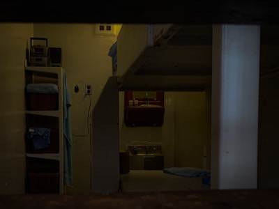 A dimly lit room with visible bunk beds, shelving units, and a washbasin. The interior appears sparse and functional, with a simple setup for personal items. The presence of folded towels and toiletries indicates a living space.