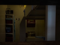 A dimly lit room with visible bunk beds, shelving units, and a washbasin. The interior appears sparse and functional, with a simple setup for personal items. The presence of folded towels and toiletries indicates a living space.