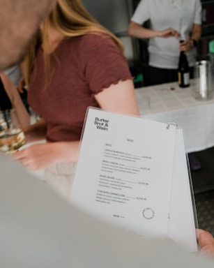 A person is holding a menu with several food items listed and priced in euros. In the background, another person is preparing a drink, which involves a bottle and a glass. The setting resembles a casual dining or catering event.