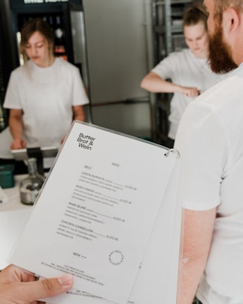 A person is holding a menu from a place called 'Butter Brot & Wein'. Two people in white shirts are visible in the background, possibly working at a counter in a food establishment.