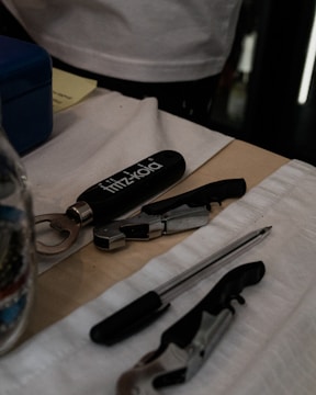 A table holds a variety of items including a fritz-kola branded bottle opener, a pen, and metallic multi-tools. The surface is covered with white fabric and a cardboard piece, while other items like a blue box and documents are partially visible.