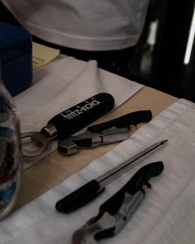 A table holds a variety of items including a fritz-kola branded bottle opener, a pen, and metallic multi-tools. The surface is covered with white fabric and a cardboard piece, while other items like a blue box and documents are partially visible.