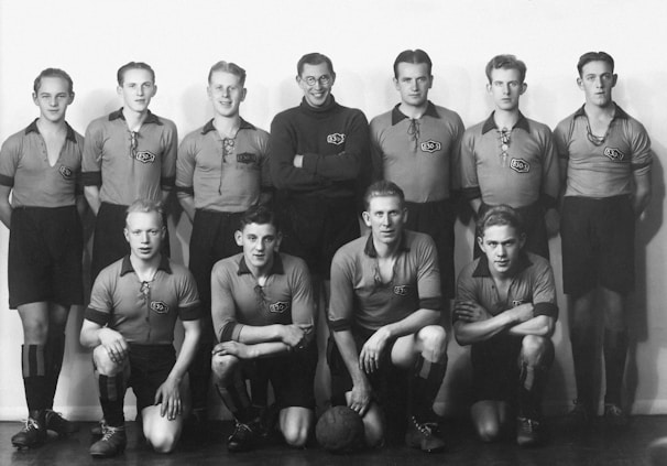 A vintage black and white photo of a men's soccer team, consisting of eleven members. The team is arranged in two rows; six standing in the back and five crouching in the front. All are wearing matching jerseys with a logo that reads '830 S' on the chest. A soccer ball is placed at the feet of the central front-row player. The setting appears to be indoors, with a plain background.