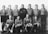 A vintage black and white photo of a men's soccer team, consisting of eleven members. The team is arranged in two rows; six standing in the back and five crouching in the front. All are wearing matching jerseys with a logo that reads '830 S' on the chest. A soccer ball is placed at the feet of the central front-row player. The setting appears to be indoors, with a plain background.
