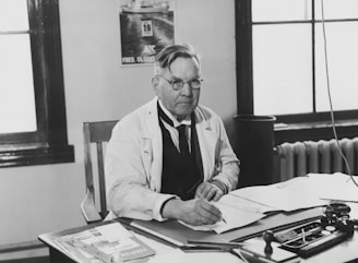 An older man wearing glasses and a white coat is seated at a desk with papers, an ink well, and writing instruments. He appears to be concentrating on his work. In the background, there is a poster and a window with panes. A radiator is visible next to the window.
