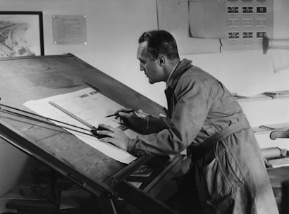 A man is working at a large drafting table, focusing intently on his work with a pencil in hand. He is wearing a long coat and appears to be engaged in architectural or engineering drawing, with various plans scattered around the table. The background includes pinned sheets and drawings, suggesting a workspace dedicated to design or planning.