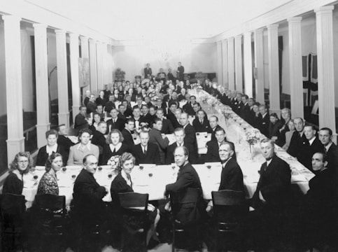 A large group of people is seated at a long, elegantly set dining table in a formal setting. Men and women, dressed in suits and dresses, are gathered together, suggesting a celebratory or formal event. The room is decorated with columns and chandeliers, providing a classic and sophisticated atmosphere. The table is adorned with floral arrangements and glassware.