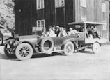 A vintage car with a connected trailer carrying several people is parked outside a wooden building. The individuals are dressed in period clothing, with hats and coats, suggesting it's from the early 20th century. The building in the background has multiple windows and is made of dark wood.
