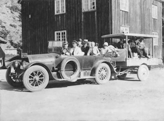 A vintage car with a connected trailer carrying several people is parked outside a wooden building. The individuals are dressed in period clothing, with hats and coats, suggesting it's from the early 20th century. The building in the background has multiple windows and is made of dark wood.