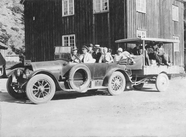 A vintage car with a connected trailer carrying several people is parked outside a wooden building. The individuals are dressed in period clothing, with hats and coats, suggesting it's from the early 20th century. The building in the background has multiple windows and is made of dark wood.