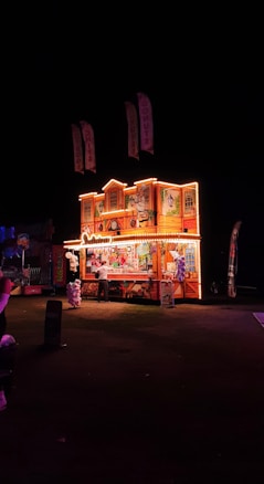 A brightly lit carnival stall at night displaying colorful illustrations and selling sweets like cotton candy and other treats. The structure is adorned with decorative lights, and a person is standing near the counter. Flags hang above the stall, and the surrounding area is dark.