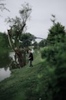 A person holding a fishing rod stands beside a river, surrounded by lush greenery. A tall tree with a slightly bent trunk is nearby, and a bridge arches over the river in the background. The scene is peaceful and overcast, indicating a cloudy day.