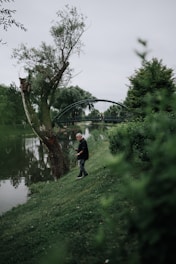 A person holding a fishing rod stands beside a river, surrounded by lush greenery. A tall tree with a slightly bent trunk is nearby, and a bridge arches over the river in the background. The scene is peaceful and overcast, indicating a cloudy day.