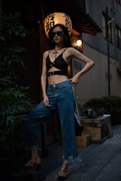 A person wearing a stylish black top and blue jeans stands confidently in an urban setting at night. They are accessorized with sunglasses and a black handbag. In the background, there is a traditional lantern with Japanese characters, giving the scene an atmospheric blend of modern and cultural elements.