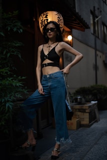 A person wearing a stylish black top and blue jeans stands confidently in an urban setting at night. They are accessorized with sunglasses and a black handbag. In the background, there is a traditional lantern with Japanese characters, giving the scene an atmospheric blend of modern and cultural elements.