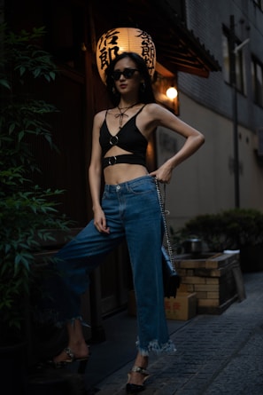 A person wearing a stylish black top and blue jeans stands confidently in an urban setting at night. They are accessorized with sunglasses and a black handbag. In the background, there is a traditional lantern with Japanese characters, giving the scene an atmospheric blend of modern and cultural elements.