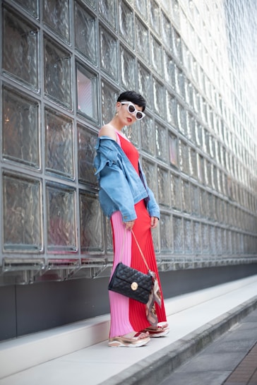 A person stands leaning against a wall made of transparent glass blocks. They are wearing a vibrant red and pink pleated dress, complemented by a faded blue denim jacket draped over their shoulders. The ensemble is accessorized with a stylish black quilted handbag and white futuristic sunglasses. The person