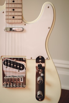 A close-up view of an electric guitar focusing on the body and the initial section of the neck. The guitar has a light cream-colored body with a glossy finish, a white pickguard, and metallic hardware including a bridge and two control knobs. The neck is made of light wood with visible grain and black dot inlays.
