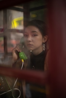 A person is inside a phone booth holding a green telephone receiver to their ear. The image is taken through the glass of the booth, giving it a slightly blurred and reflective effect. The person has dark hair and is wearing large earrings.