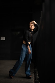 A person stands in a dimly lit corridor, wearing a black long-sleeve crop top and loose-fitting blue jeans. Their head is tilted with one hand resting on their head, and they are leaning against a black railing. The expression appears contemplative, adding to the moody atmosphere.