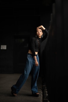 A person stands in a dimly lit corridor, wearing a black long-sleeve crop top and loose-fitting blue jeans. Their head is tilted with one hand resting on their head, and they are leaning against a black railing. The expression appears contemplative, adding to the moody atmosphere.