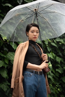A person stands confidently under a transparent umbrella, surrounded by lush green foliage. They are wearing a black top, blue jeans, and a tan coat draped over their shoulders, accessorized with statement earrings and a chain across their body. The umbrella is speckled with raindrops, suggesting a light rain.