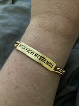 A close-up view of a wrist wearing a gold bracelet with an engraved message that reads, 'Bitch, you're my soulmate.' The skin tone is light, and the background is blurred, focusing attention on the bracelet.