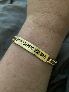 A close-up view of a wrist wearing a gold bracelet with an engraved message that reads, 'Bitch, you're my soulmate.' The skin tone is light, and the background is blurred, focusing attention on the bracelet.