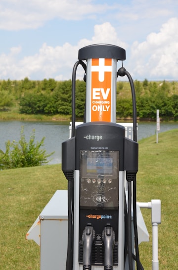 An electric vehicle (EV) charging station is situated in an outdoor, grassy area with trees in the background. The station features a ChargePoint unit with two charging plugs and an orange sign indicating 'EV Charging Only'. A small body of water is also visible behind the station.