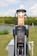 An electric vehicle (EV) charging station is situated in an outdoor, grassy area with trees in the background. The station features a ChargePoint unit with two charging plugs and an orange sign indicating 'EV Charging Only'. A small body of water is also visible behind the station.
