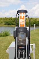 An electric vehicle (EV) charging station is situated in an outdoor, grassy area with trees in the background. The station features a ChargePoint unit with two charging plugs and an orange sign indicating 'EV Charging Only'. A small body of water is also visible behind the station.