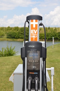 An electric vehicle (EV) charging station is situated in an outdoor, grassy area with trees in the background. The station features a ChargePoint unit with two charging plugs and an orange sign indicating 'EV Charging Only'. A small body of water is also visible behind the station.