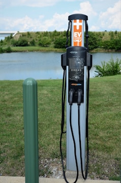 An electric vehicle charging station is situated outdoors near a body of water with green grass and some bushes in the background. The station has two charging cables and connectors hanging neatly. A green pole is seen to the left of the charger, possibly marking or protecting the charging area.