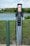 An electric vehicle charging station is situated outdoors near a body of water with green grass and some bushes in the background. The station has two charging cables and connectors hanging neatly. A green pole is seen to the left of the charger, possibly marking or protecting the charging area.
