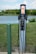 An electric vehicle charging station is situated outdoors near a body of water with green grass and some bushes in the background. The station has two charging cables and connectors hanging neatly. A green pole is seen to the left of the charger, possibly marking or protecting the charging area.