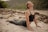A person is performing a yoga pose on a sandy ground near a river with rocks and green foliage in the background. They are wearing a black sports bra and green leggings, extending their body forward and stretching upward while keeping their eyes closed.